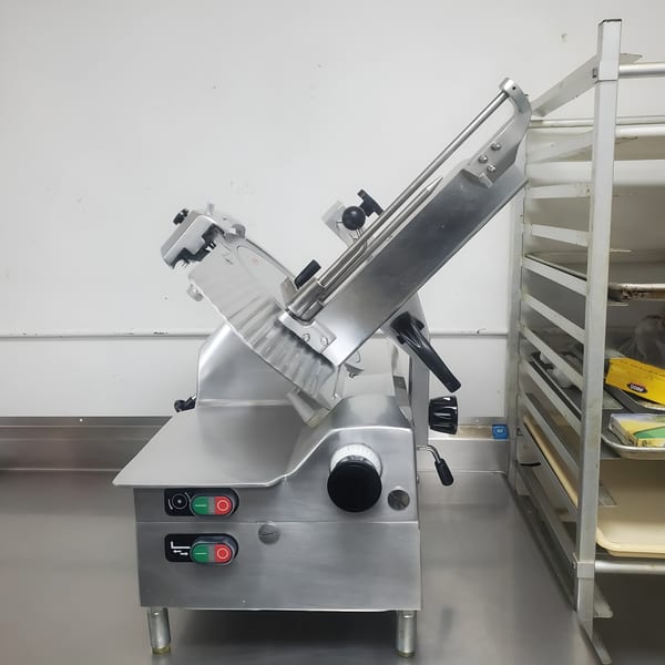 Stainless steel commercial meat slicer with red and green buttons on a metal counter next to a baking rack.