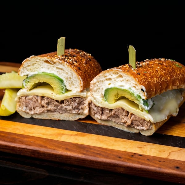Two halves of a hearty sandwich with meat, cheese, avocado, and sauce, served with a pickle.