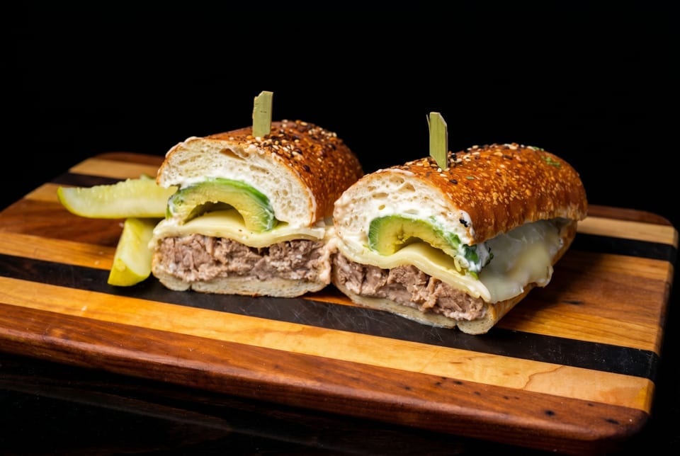 Two halves of a hearty sandwich with meat, cheese, avocado, and sauce, served with a pickle.