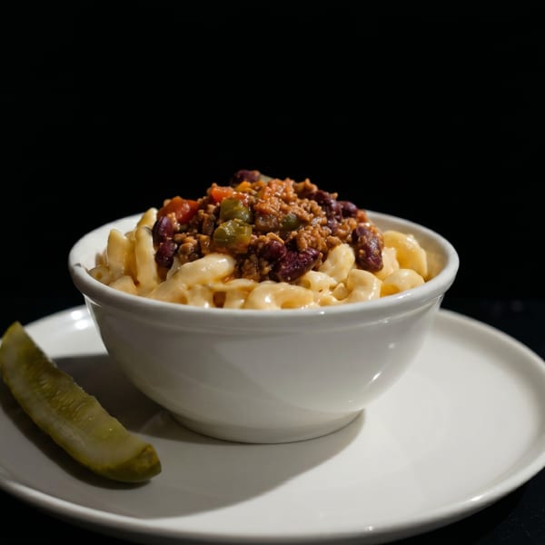 A white bowl of macaroni and cheese topped with chili, served with a pickle spear.