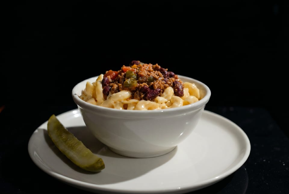 A white bowl of macaroni and cheese topped with chili, served with a pickle spear.