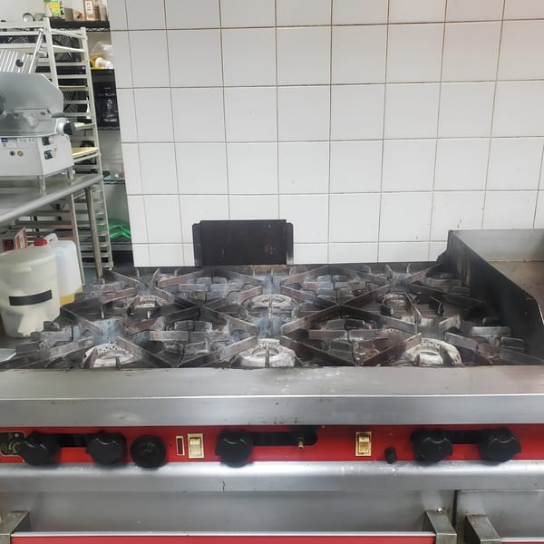 A commercial kitchen gas range with six burners and a flat top, set against white tiled walls.