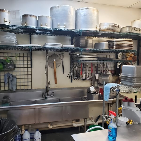 A commercial kitchen with metal shelving, large pots, a triple sink, and various cooking utensils.