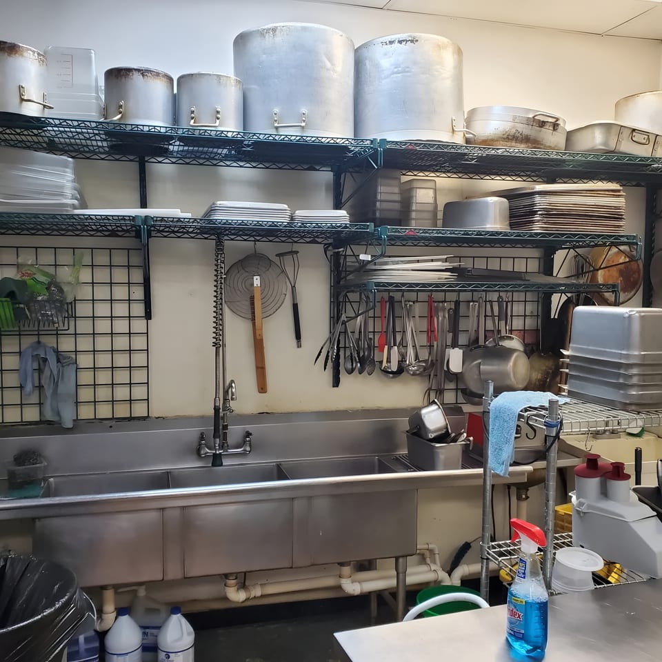 A commercial kitchen with metal shelving, large pots, a triple sink, and various cooking utensils.