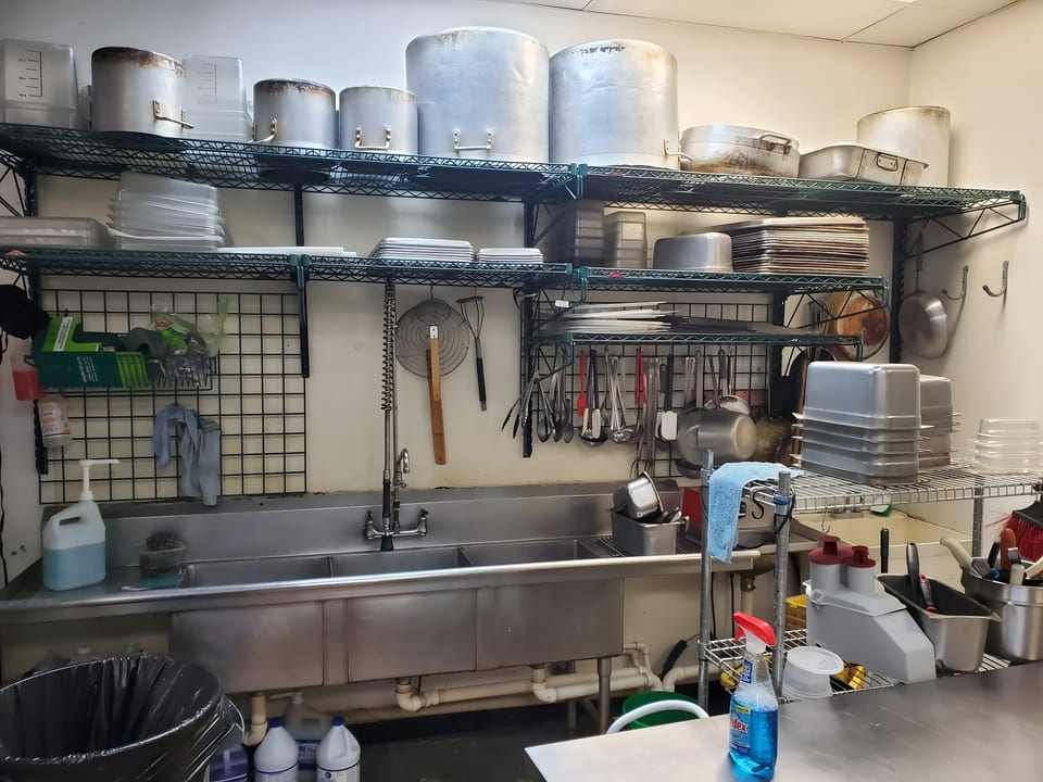 A commercial kitchen with metal shelving, large pots, a triple sink, and various cooking utensils.