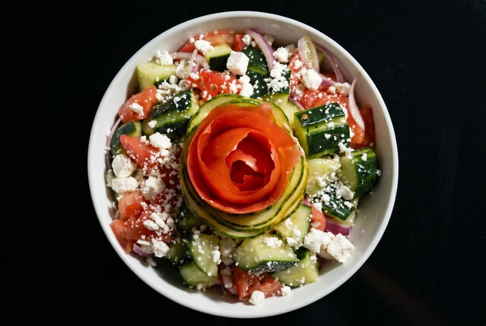 A vibrant Greek salad with cucumbers, tomatoes, red onion, feta, and a cucumber-tomato rose garnish.