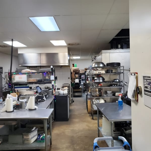 A commercial kitchen with stainless steel tables, shelving, and cooking equipment under fluorescent lights.