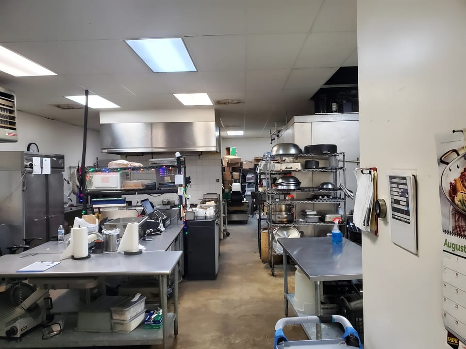 A commercial kitchen with stainless steel tables, shelving, and cooking equipment under fluorescent lights.