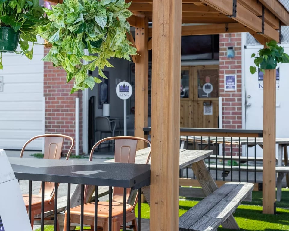 Outdoor patio with hanging plants, metal chairs, and wooden picnic tables on artificial grass.