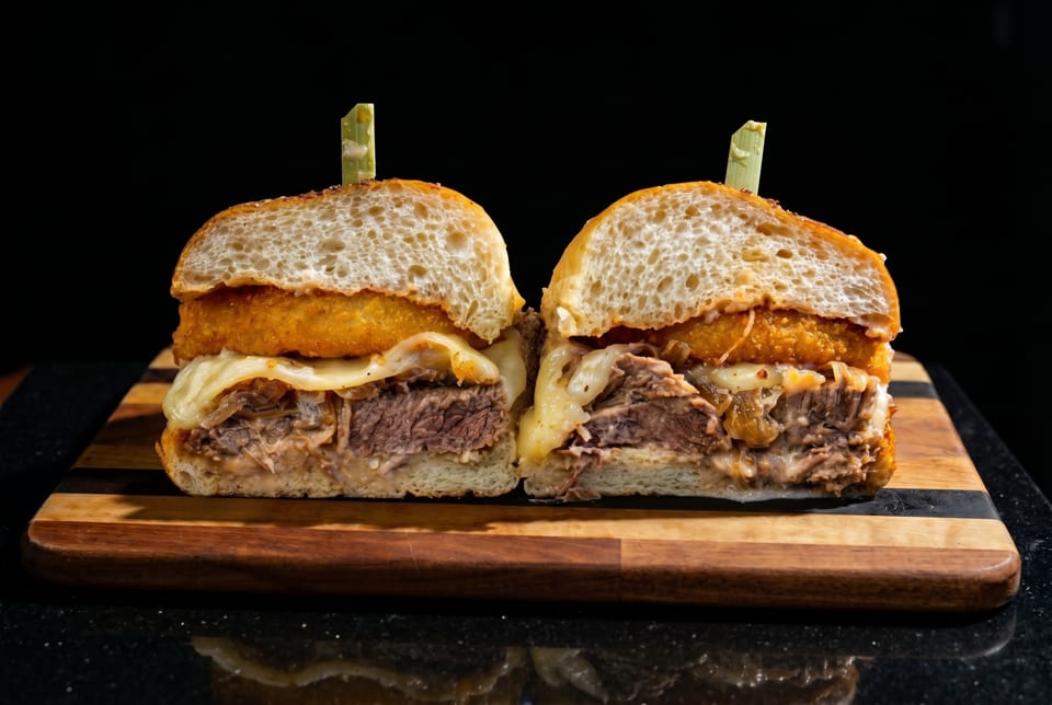 A large sandwich, cut in half, revealing beef, melted cheese, and a fried patty on a wooden board.