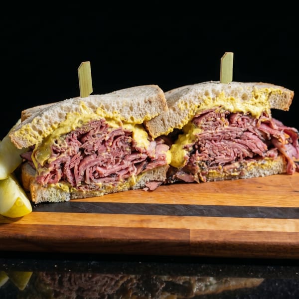 Pastrami sandwich with mustard and pickles on a wooden board against a dark background.