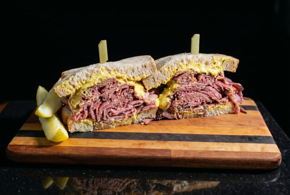 Pastrami sandwich with mustard and pickles on a wooden board against a dark background.