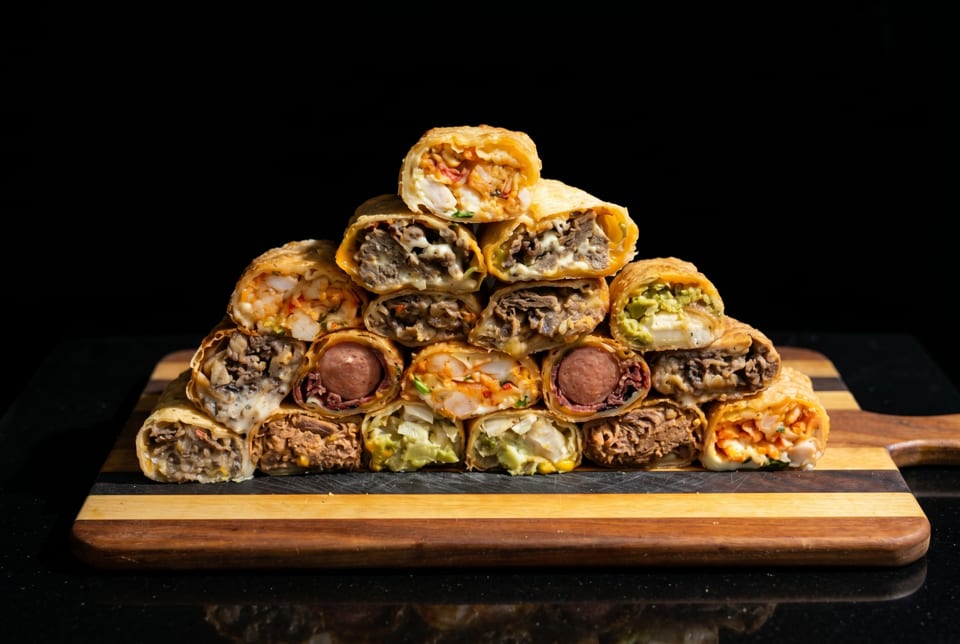 A pyramid of various crispy, cut-open burritos with diverse fillings on a wooden board.