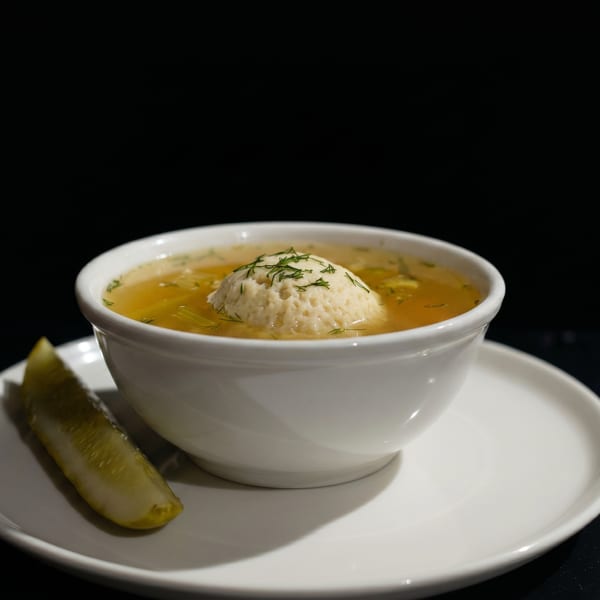 Matzo ball soup with dill and a pickle spear on a white plate against a dark background.