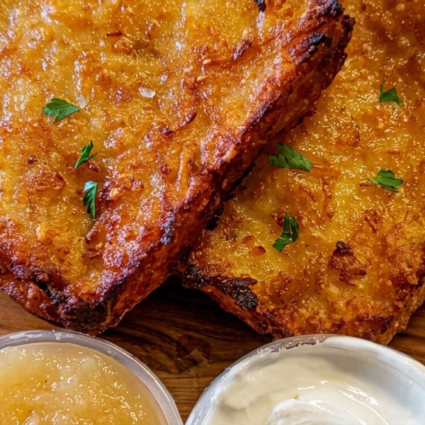 Two golden-brown potato latkes with parsley, applesauce, and sour cream on a wooden board.